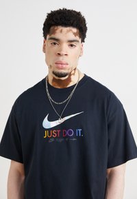Juodas medvilninis marškinėliai su užrašu "TIESIOG DARYK" vaivorykštės spalvomis ir atspindinčiu Nike logotipu. Modelis dėvi sidabrinius grandininius vėrinius.