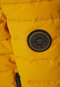 Yellow padded jacket with vertical ribbed texture. Features a black circular logo patch with white and gold accents on the upper arm.