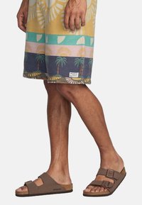 Sandali Birkenstock marroni con due fibbie; soletta in sughero testurizzata; indossati con pantaloni corti a fantasia multicolore che presentano motivi di palme e sole.