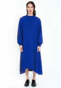 Mittellanges blaues Kleid mit langen Ballonärmeln, hohem Kragen und leicht gebogenem Saum. Aus gleichem Stoff gefertigt und mit schwarzen Schuhen kombiniert.