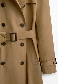 Beige trench coat with a double-breasted design, black buttons, a belt at the waist, and side pockets. Smooth fabric with a slight sheen.