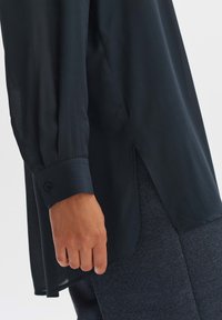 Schwarzes Langarmshirt mit hohem Schlitz und geknöpften Ärmeln, aus leichtem Stoff gefertigt, der eine glatte Textur aufweist und keine sichtbaren Muster hat.