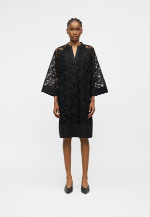 Žena stojaca v čiernych čipkovaných šatách po kolená so širokými rukávmi a výstrihom do V, doplnená čiernymi špicatými topánkami na jednoduchom pozadí.