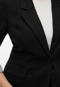 Zwarte blazer met een gefacetteerde revers, enkele knoopsluiting en een voorvak. Gedragen over een witte, gestructureerde top.