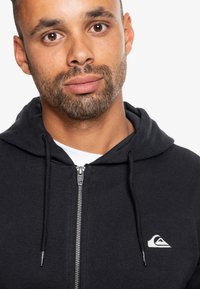 Čierna mikina na zips s kapucňou a bielym logom na ľavej hrudi. Materiál má jemnú textúru a neformálny dizajn.