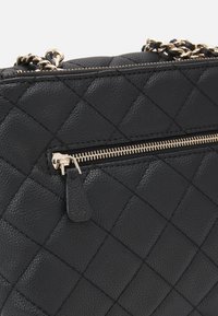 Sac à main en cuir matelassé noir avec quincaillerie dorée, une poche zippée et des détails en chaîne sur la bandoulière. Surface texturée avec des motifs en diamant.