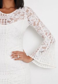 Robe en dentelle blanche avec un corsage ajusté et des manches en forme de cloche. Le tissu présente des motifs floraux et une finition texturée, avec des sections transparentes.