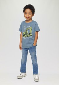 Blauwe katoenen T-shirt met een gele graafmachineafbeelding en witte tekst, gecombineerd met lichtblauwe spijkerbroek en witte sneakers met grijze accenten.