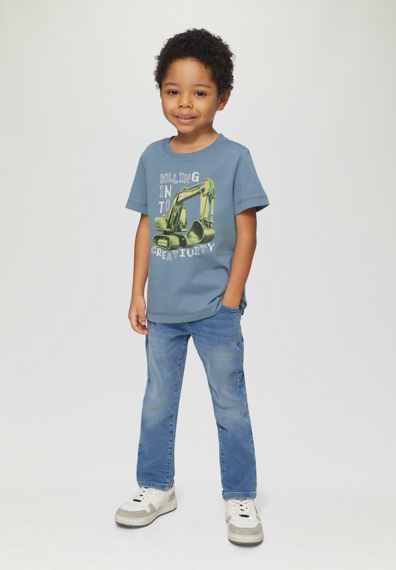 Blauwe katoenen T-shirt met een gele graafmachineafbeelding en witte tekst, gecombineerd met lichtblauwe spijkerbroek en witte sneakers met grijze accenten.