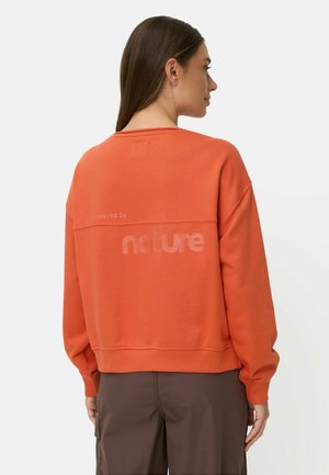 Kvinde med langt brunt hår bærer orange sweatshirt med teksten "nature" på ryggen og mørkebrune bukser, vendt væk fra hvid baggrund.