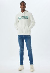Sudadera blanca con "Departamento Atlético de California" impreso en verde, combinada con pantalones vaqueros azules y zapatillas de color claro. El modelo está de pie.
