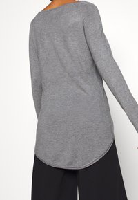 Maglia a maniche lunghe grigia in maglia con orlo arrotondato, caratterizzata da una sottile texture e un scollo ampio. Indossata con pantaloni neri larghi.