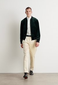 Chaqueta bomber de terciopelo verde sobre una camisa blanca, combinada con pantalones beiges y zapatos grises. El conjunto presenta un diseño elegante y moderno.
