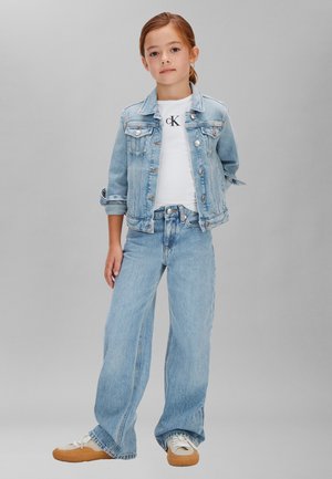 Fiatal lány áll, kezei mellett, világoskék farmerdzsekit és farmert, fehér pólót, valamint fehér, barna talpú sportcipőt visel.