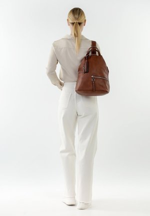 Mochila de cuero marrón con forma redondeada, bolsillo frontal con cremallera y un detalle de borlas en un lateral, llevada sobre una camisa blanca y pantalones.