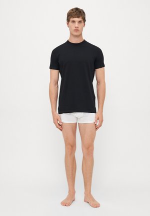 Jeune homme debout pieds nus sur un fond blanc, portant un T-shirt noir uni et un boxer blanc.