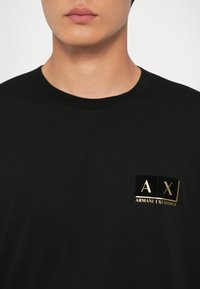 Černé tričko s krátkým rukávem vyrobené z bavlny, s logem vyšitým ve zlaté barvě: "AX" a "ARMANI EXCHANGE" na levém hrudníku.