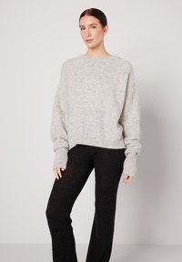 Tumejuustega naine, kes kannab lahtist helehalli kampsunit ja kehakuvi järgivaid musti pükse, seisab ühevärvilise heleda tausta ees.