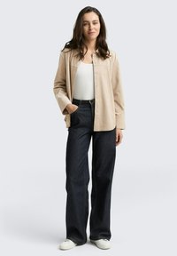 CHAQUETA BEIGE DE BOTONES SOBRE UNA CAMISETA BLANCA, COMBINADA CON PANTALONES DE PIERNA ANCHA OSCUROS Y ZAPATILLAS BLANCAS; TEJIDO SUAVE, CORTE RECTO Y COSTURAS SUTILS.