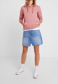 Woman wearing a pink hoodie, light blue denim skirt with frayed hem, white socks, and white high-top sneakers standing with hands in pockets.