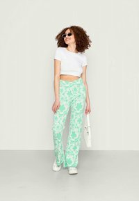 Calças verdes menta largas com um padrão floral, combinadas com um top cropped branco e ténis brancos, acompanhadas de óculos de sol pretos e uma bolsa.