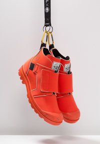 Bottes orange vif avec semelles épaisses et larges sangles en velcro, suspendues par des mousquetons jaunes attachés à une sangle noire.