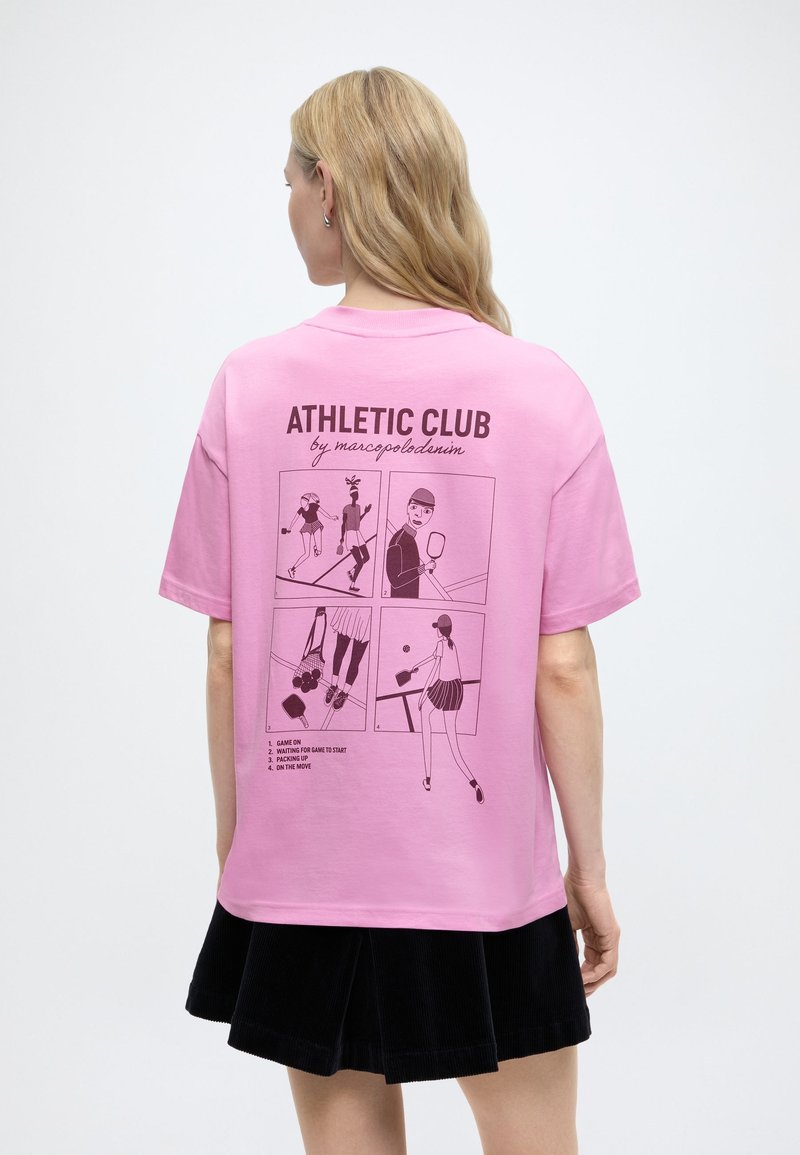 Mujer con cabello rubio largo, vistiendo una camiseta rosa con ilustraciones de tenis estilo cómic en negro y texto en la espalda que dice "ATHLETIC CLUB".