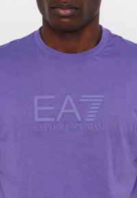 Violet medvilnės marškinėliai su priekyje esančiu grafiniu logotipu toniniu spaudiniu. Apvalus iškirpimas, trumpomis rankovėmis, minkšta tekstūra ir nevaržantis siluetas.