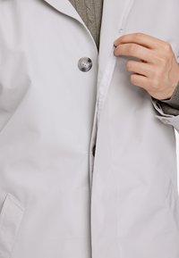 Giacca impermeabile grigio chiaro con colletto a risvolto, dotata di chiusura a bottoni e tasche laterali, che presenta un tessuto liscio e testurizzato.