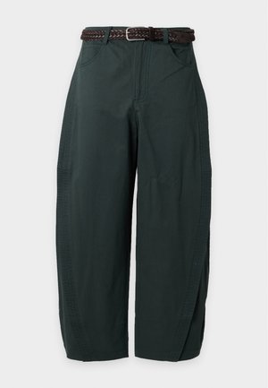 Joven mujer con cabello largo y oscuro lleva un top de punto a rayas crema, pantalones anchos verde oscuro con cinturón y zapatos planos negros, de pie con una mano en el bolsillo.