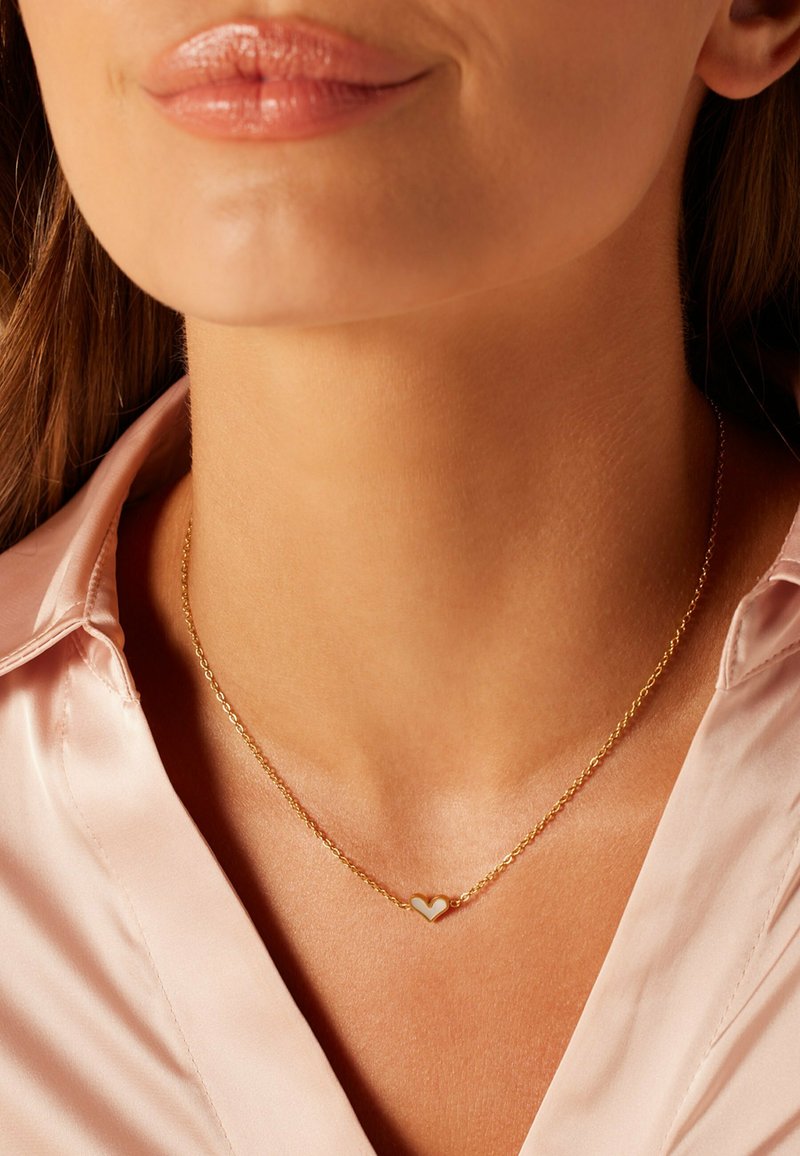 Gouden ketting met een kleine hartjeshanger. Het hart heeft een witte emaille inset, rustend op een gladde, blote hals.