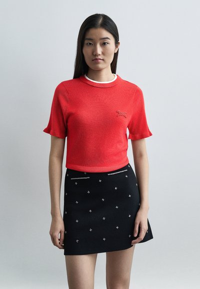 Jeune femme aux longs cheveux bruns portant un haut rouge à manches courtes avec un logo de cheval et une mini-jupe noire ornée de clous argentés.