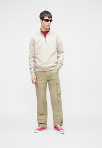 Junger Mann, der schwarze Sonnenbrille, einen beigen Sweatshirt mit viertel Reißverschluss, khakifarbene Cargo-Hosen und rote Sneakers trägt und vor einem weißen Hintergrund steht.