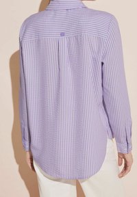 Lila randig skjorta med knapp-down krage, långa ärmar och rundad nederkant. Texturerad tyg med ljusare lila och vita vertikala ränder.