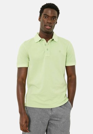 Junger Mann steht vor einem einfarbigen Hintergrund, trägt ein hellgrünes Polohemd mit kurzen Ärmeln und graue Jeans, die eine Hand in der Tasche hat.