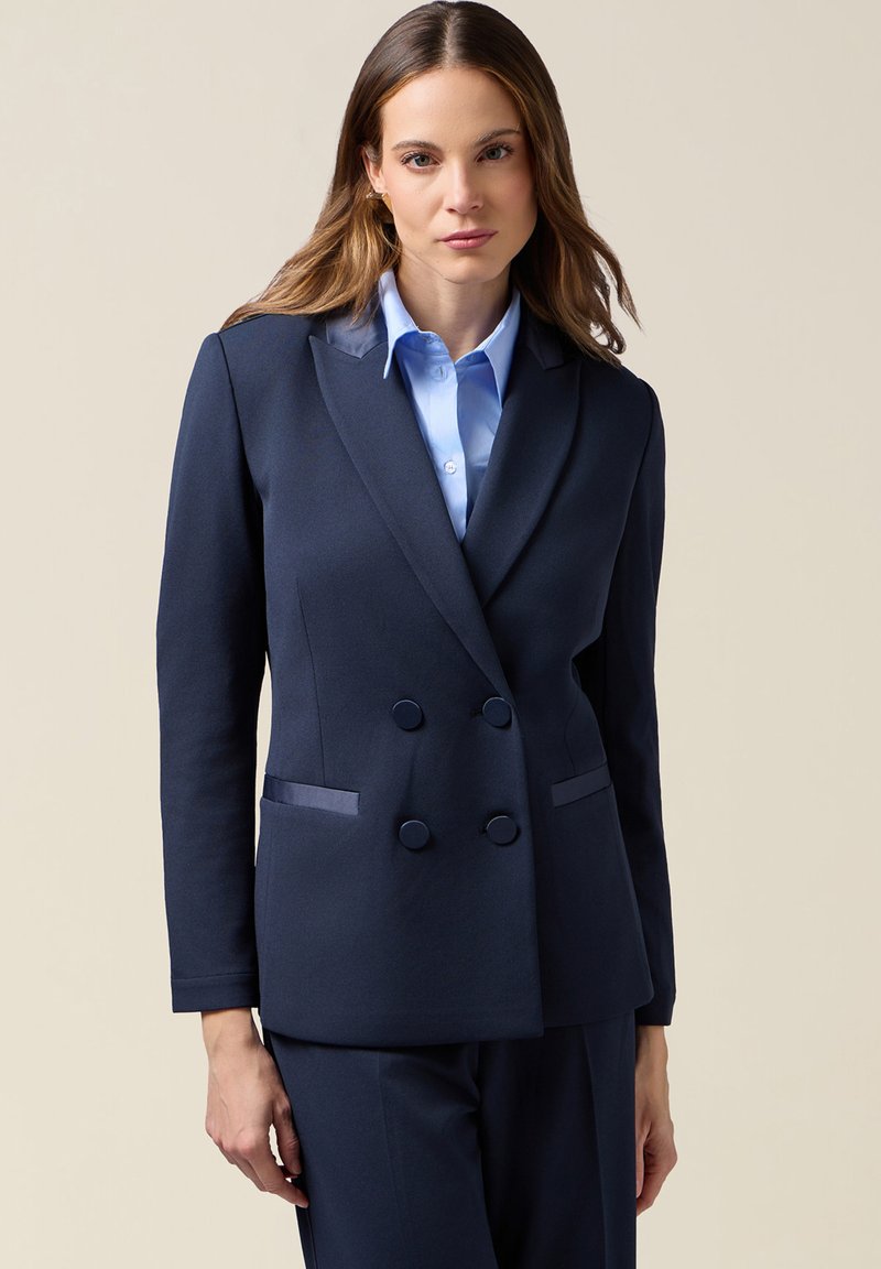 Blazer doppiopetto blu navy con due tasche frontali, bottoni arrotondati e una vestibilità leggermente sagomata, abbinato a una camicia celeste con colletto.