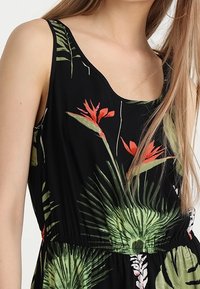 Svart ärmlös klänning med tropiska blommönster i grönt och rött, med elastisk midja och mjuk tygtextur.