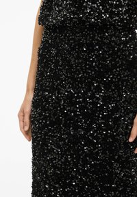 Robe noire à paillettes présentant une surface texturée avec des paillettes rondes superposées. Le tissu est lisse, brillant et moulant.