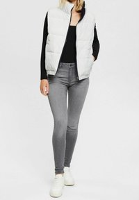 Light gray puffer vest, zippered, with a high collar, paired with form-fitting gray jeans and white sneakers. Simple, functional design.