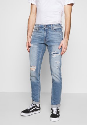 Mann trägt hellblaue zerrissene Jeans, schwarz-weiße Turnschuhe und ein weißes kurzärmeliges Hemd, steht vor einem schlichten Hintergrund.