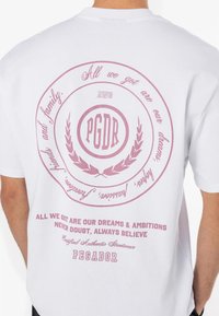 Persona vistiendo una camiseta blanca con un diseño de texto circular rosa que presenta "PGDR" y frases motivacionales en la parte trasera.