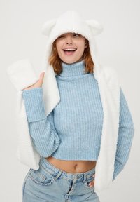 Niebieski sweter robiony na drutach, krótki fason, z golfem. Biały pluszowy szalik z kapturem i uszami misia. Modelka prezentuje rękawiczki, uśmiechając się, w dżinsowych spodniach.