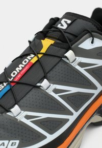 Γκρι αθλητικό παπούτσι με μαύρα κορδόνια. Διαθέτει πολύχρωμες λεπτομέρειες σε κίτρινο, κόκκινο και μπλε. Υφασμάτινο πάνω μέρος με λευκές αντανακλαστικές λεπτομέρειες, πορτοκαλί σόλα.