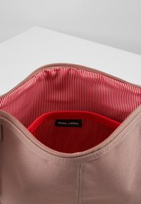 Borsa in tessuto marrone chiaro con apertura a zip, dotata di un interno a righe rosse, tasca in rete e etichetta "BENVENUTO A HERSCHEL".