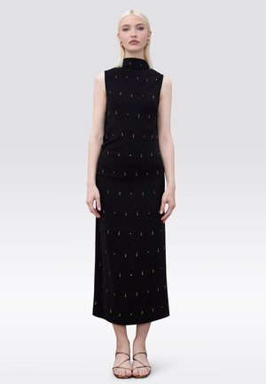 Mujer con cabello rubio liso, vestida con un vestido midi negro sin mangas adornado con puntos dorados y sandalias doradas de tiras, de pie inmóvil.