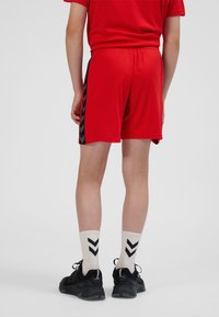 Rode sportkorte broek met zwarte chevronstrepen aan de zijkant, gedragen met witte sokken met een zwart pijlenpatroon en zwarte sneakers.