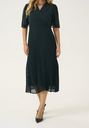 Femme en robe midi plissée vert foncé avec manches courtes transparentes et escarpins noirs à bout pointu, debout devant un fond clair uni.