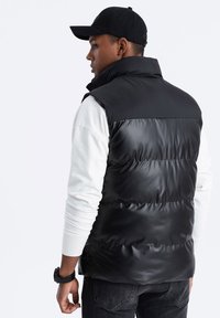 Schwarze Weste aus Puffer-Material mit hohem Kragen und glatter Textur. Über einem weißen Langarmshirt getragen. Signaturkappe und dunkle Jeans sichtbar.