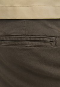 Gros plan d'une poche de pantalon en tissu marron avec détail de couture et ceinture beige au-dessus.