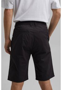 Homme portant un short noir aux genoux et un t-shirt blanc, vu de dos sur un fond neutre.
